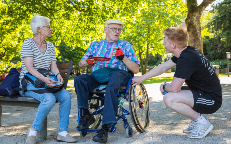 Drei Personen im Park. Eine ältere Dame sitzt auf einer Bank. Ein älterer Mann im Rollstuhl. Daneben hockt ein jüngerer Mann und die drei unterhalten sich.