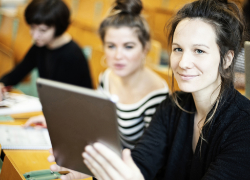 Drei junge Frauen sitzen in einem Hörsaal. Die Frau im Vordergrund hat ein Tablet in der Hand und schaut in die Kamera.