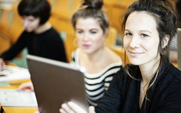 Drei junge Frauen sitzen in einem Hörsaal. Die Frau im Vordergrund hat ein Tablet in der Hand und schaut in die Kamera.