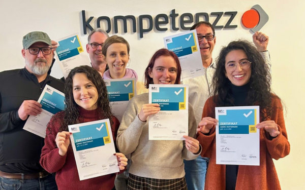 Sieben Mitarbeitende stehen vor dem Logo des Kompetenzzentrums und halten lächelnd ihr Digitalführerschein-Zertifikat in die Kamera.