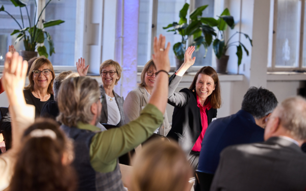 Mehrere Personen sitzen in einem Veranstaltungssaal. Einige drehen sich um und lächeln, einige heben die Hand.