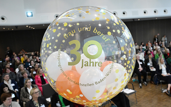 Ein Ballon mit den Worten 30 Jahre Frau und Beruf, Landesprogramm Kontaktstelle Baden-Württemberg in goldener Schrift. Im Hintergrund ist ein Saal voller Menschen zu sehen.