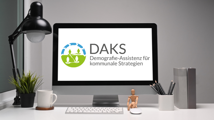 Ein Schreibtisch mit Utensilien und einem Bildschirm auf dem das Logo der "Demografie-Assistenz für kommunale Strategien (DAKS)" zu sehen ist.