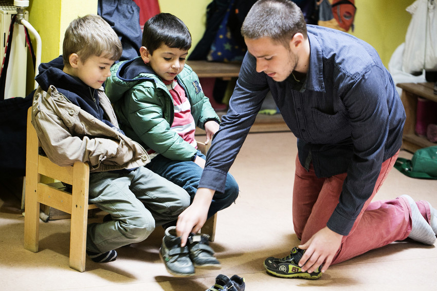 Ein junger Mann hilft zwei Jungen im Kindergartenalter beim Schuhe anziehen.