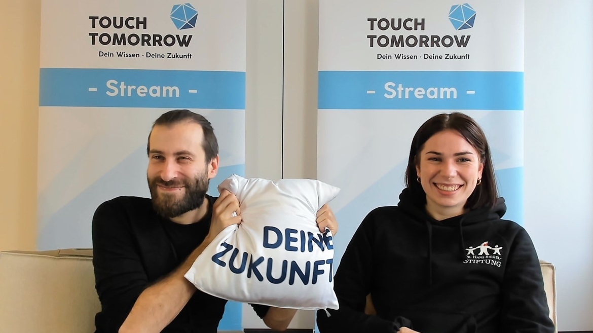 Ein junger Mann und eine junge Frau sitzen vor einem Banner des Touch-Tomorrow-Streams und halten lächelnd ein Kissen in die Kamera, auf dem "Deine Zukunft" steht.