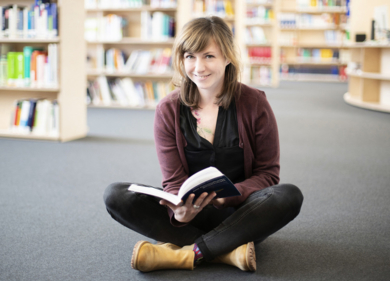 Junge Frau mit einem Buch in der Hand sitzt auf dem Boden einer Bibliothek
