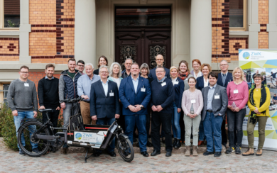 Eine Gruppe von ca. 20 Erwachsenen lächelt in die Kamera. Am Rand das Roll-Up der ZWK. Teilnehmende des Regionalen Werkstatttreffens in Barleben.